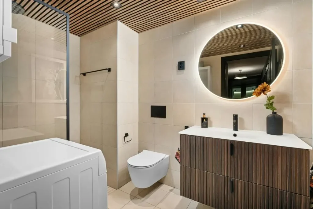 Modern small bathroom featuring a sleek design with a round illuminated mirror, wooden accents, a wall-mounted toilet, and a stylish vanity, emphasizing functionality and aesthetics in compact spaces.